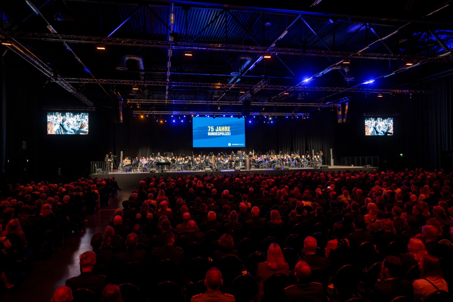 Volle Halle mit Eventbeleuchtung, mit froßem Orchester auf der Bühne.